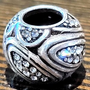 May include: A silver-toned bead with a central hole, adorned with clear, sparkling crystals. The bead features a swirling, textured design, creating a decorative and elegant appearance. The bead is likely for jewelry making.