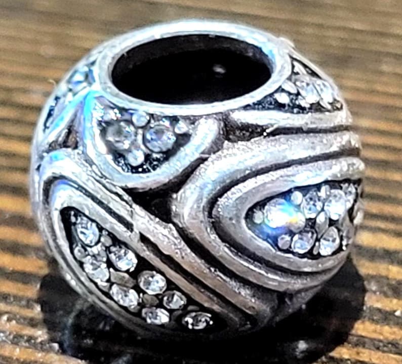 May include: A silver-toned bead with a central hole, adorned with clear, sparkling crystals. The bead features a swirling, textured design, creating a decorative and elegant appearance. The bead is likely for jewelry making.