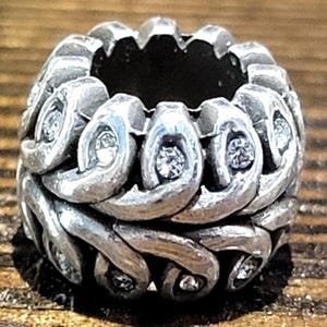 May include: A silver-toned ring with an intricate, woven design. The ring features small, clear gemstones set within the woven pattern. The ring's design is detailed and appears to be made of metal.