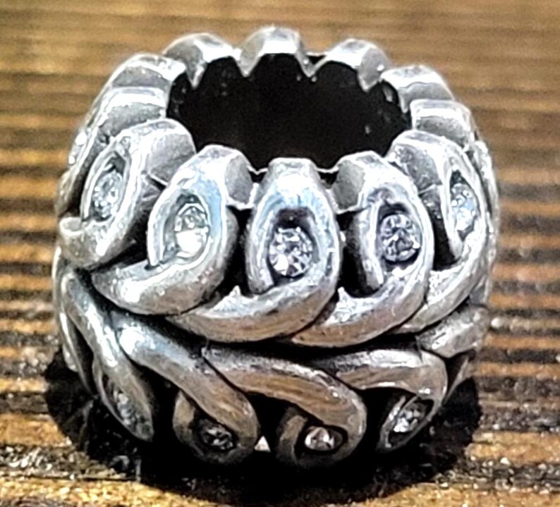 May include: A silver-toned ring with an intricate, woven design. The ring features small, clear gemstones set within the woven pattern. The ring's design is detailed and appears to be made of metal.