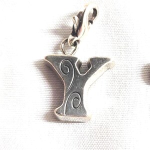 May include: Silver-toned metal charm in the shape of the letter "Y" with decorative swirls. The charm has a clasp for easy attachment to a bracelet or necklace. Other letter charms are visible.