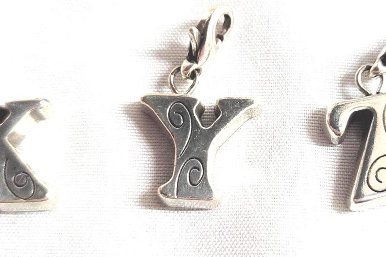 May include: Silver-toned metal charm in the shape of the letter "Y" with decorative swirls. The charm has a clasp for easy attachment to a bracelet or necklace. Other letter charms are visible.