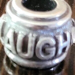 May include: A silver charm with the word "LAUGH" embossed around the center. The charm has a cylindrical shape with a dark interior and decorative detailing. The letters are in a bold, rounded font.