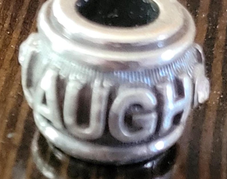 May include: A silver charm with the word "LAUGH" embossed around the center. The charm has a cylindrical shape with a dark interior and decorative detailing. The letters are in a bold, rounded font.
