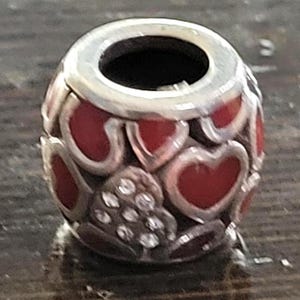 May include: A silver charm bead with a red heart design. The bead has a cylindrical shape with a silver band at the top. The red hearts are interspersed with clear, sparkling accents.