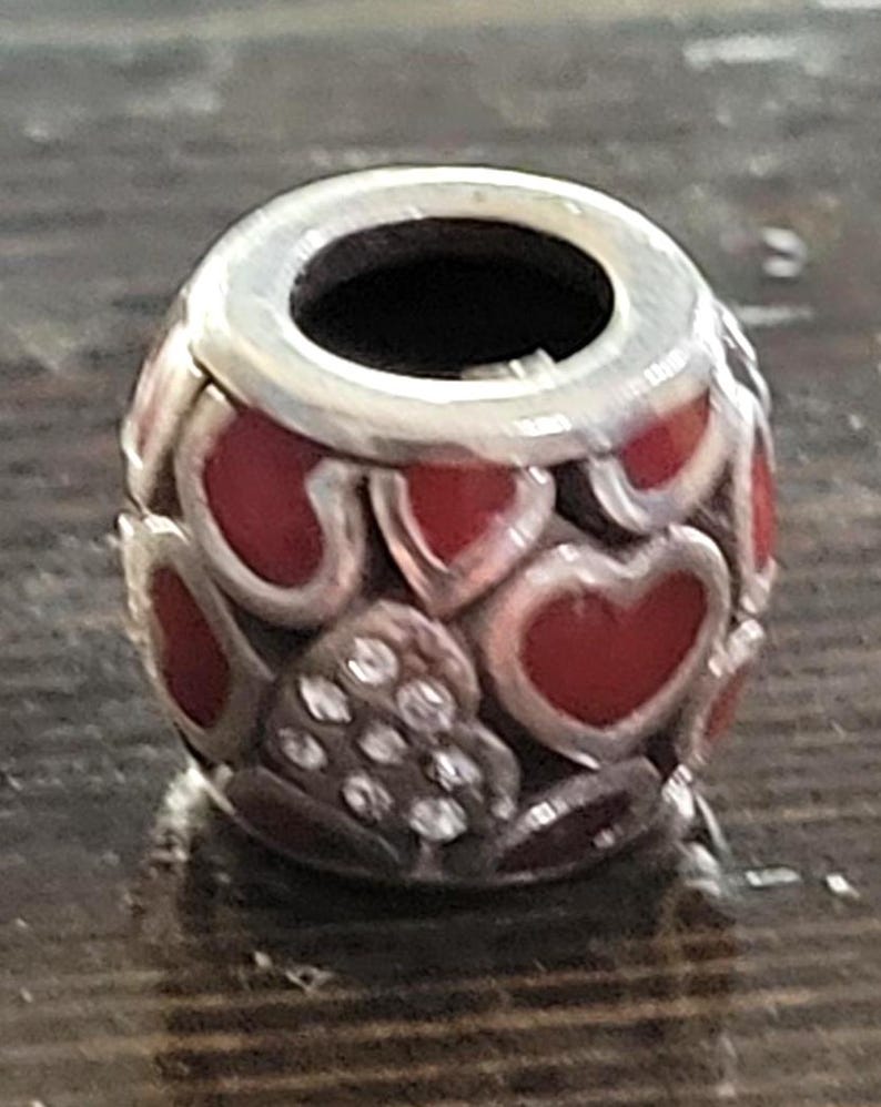 May include: A silver charm bead with a red heart design. The bead has a cylindrical shape with a silver band at the top. The red hearts are interspersed with clear, sparkling accents.