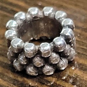 May include: A silver-colored ring with a textured surface. The ring is made of small, round beads arranged in a circular pattern. The beads have a slightly irregular shape, giving the ring a unique, handcrafted look.