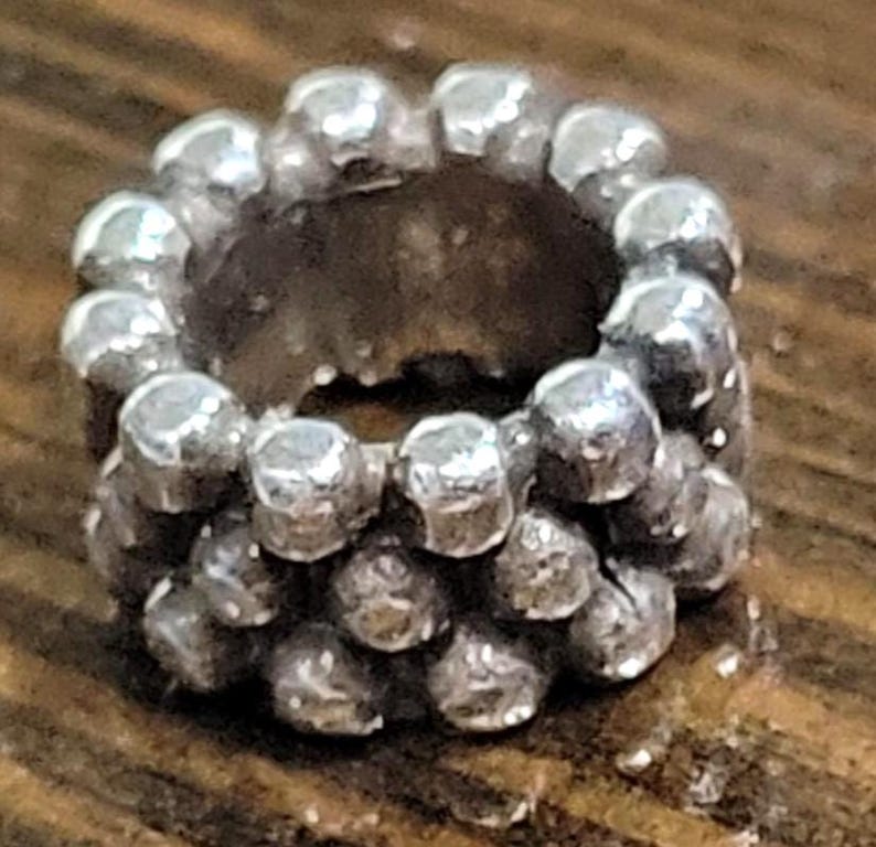 May include: A silver-colored ring with a textured surface. The ring is made of small, round beads arranged in a circular pattern. The beads have a slightly irregular shape, giving the ring a unique, handcrafted look.