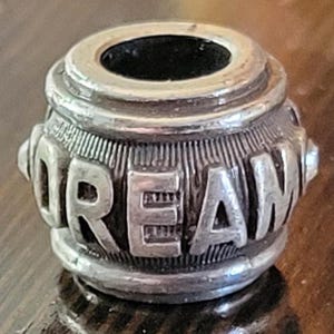 May include: A silver-toned bead with the word "DREAM" embossed around its cylindrical body. The bead has a hole running through the center and is likely used for jewelry making or as a decorative element.