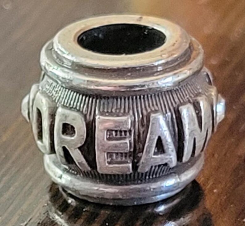 May include: A silver-toned bead with the word "DREAM" embossed around its cylindrical body. The bead has a hole running through the center and is likely used for jewelry making or as a decorative element.