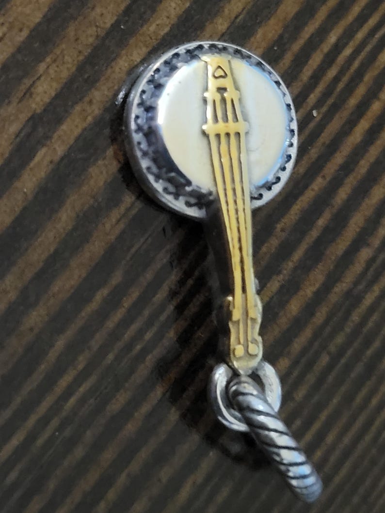 Puede incluir: Un dije en forma de banjo con un cuerpo de metal plateado y un dise&ntilde;o de banjo dorado. El dije tiene una cara blanca y crema con un borde moteado negro. El dije est&aacute; unido a un lazo de metal retorcido plateado.