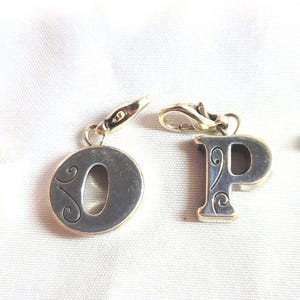 May include: Four silver-toned letter charms: N, O, P, and Q. Each charm has a decorative, swirling design and a clasp for attaching to a bracelet or necklace. The letters are displayed on a white surface.