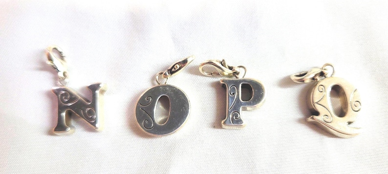 May include: Four silver-toned letter charms: N, O, P, and Q. Each charm has a decorative, swirling design and a clasp for attaching to a bracelet or necklace. The letters are displayed on a white surface.