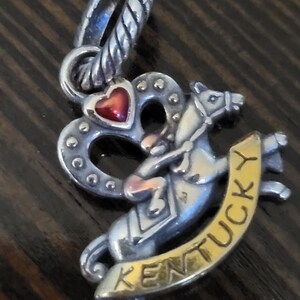Puede incluir: Charm de plata con un coraz&oacute;n rojo y un caballo con un jinete. El charm tiene la palabra "Kentucky" en una pancarta dorada. Dise&ntilde;ado para sujetar a una pulsera o collar.