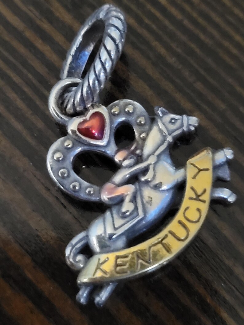 Puede incluir: Charm de plata con un coraz&oacute;n rojo y un caballo con un jinete. El charm tiene la palabra "Kentucky" en una pancarta dorada. Dise&ntilde;ado para sujetar a una pulsera o collar.