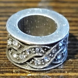 May include: A silver-toned ring with a decorative band. The band features a swirling design adorned with small, clear gemstones. The ring has a circular shape with a hollow center, and is likely a bead or charm.