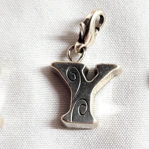 May include: Three silver-toned alphabet charms: X, Y, and Z. Each charm features a decorative swirl design and a clasp for attachment. The charms are displayed on a white, textured surface.