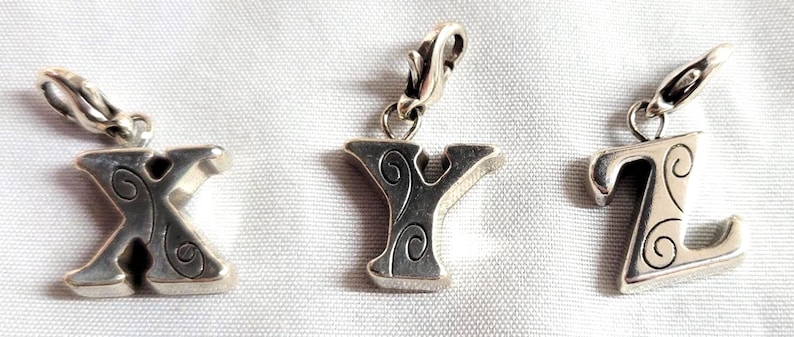 May include: Three silver-toned alphabet charms: X, Y, and Z. Each charm features a decorative swirl design and a clasp for attachment. The charms are displayed on a white, textured surface.