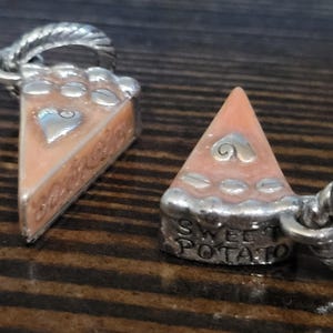 Puede incluir: Dos colgantes plateados con forma de porciones de tarta de boniato. Cada porci&oacute;n tiene un relleno naranja claro, una corteza plateada y un dise&ntilde;o de coraz&oacute;n. Un colgante tiene las palabras "Sweet Potato" en la base.