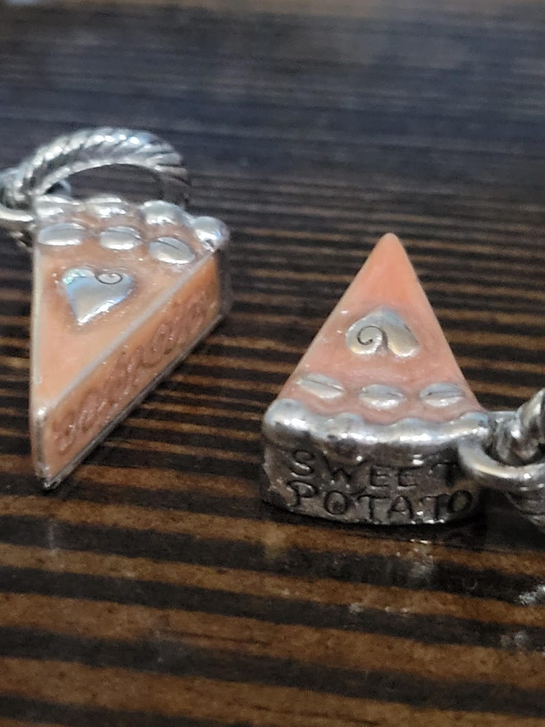 Puede incluir: Dos colgantes plateados con forma de porciones de tarta de boniato. Cada porci&oacute;n tiene un relleno naranja claro, una corteza plateada y un dise&ntilde;o de coraz&oacute;n. Un colgante tiene las palabras "Sweet Potato" en la base.