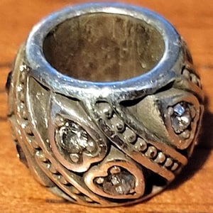 May include: Silver-toned bead with a central hole, featuring heart-shaped and dotted patterns. The bead is adorned with small, clear gemstones. The design is intricate and detailed, with a vintage or antique aesthetic.