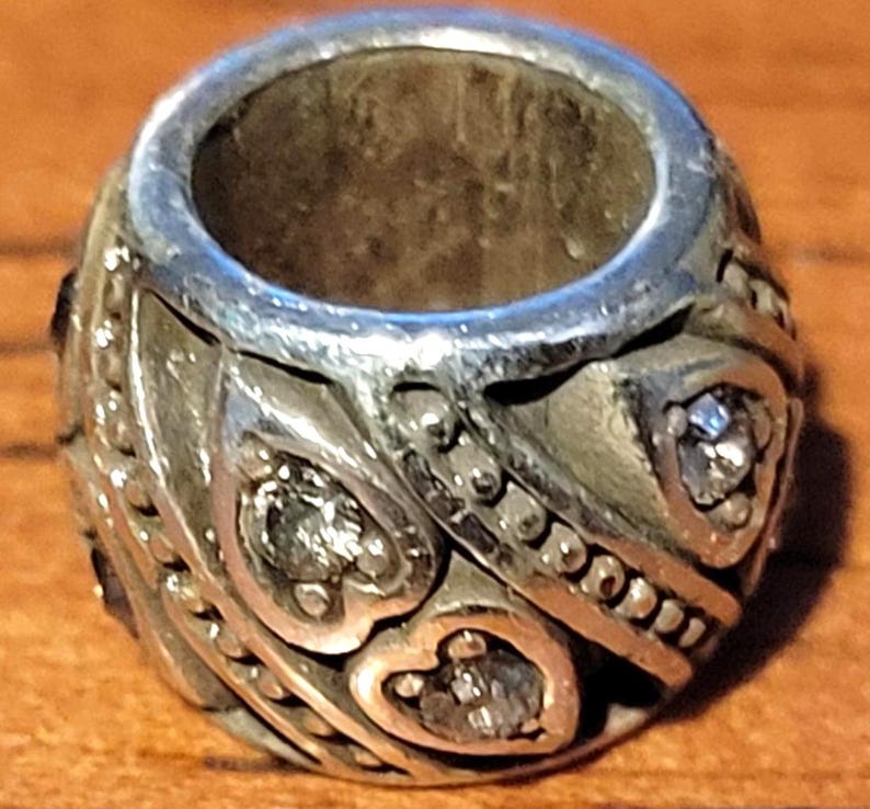 May include: Silver-toned bead with a central hole, featuring heart-shaped and dotted patterns. The bead is adorned with small, clear gemstones. The design is intricate and detailed, with a vintage or antique aesthetic.