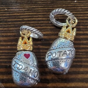Puede incluir: Dos colgantes plateados con forma de patata y coronas doradas. Uno tiene grabado "IDAHO" con un coraz&oacute;n rojo, y el otro "POTATO". Ambos colgantes tienen un anillo plateado en la parte superior.