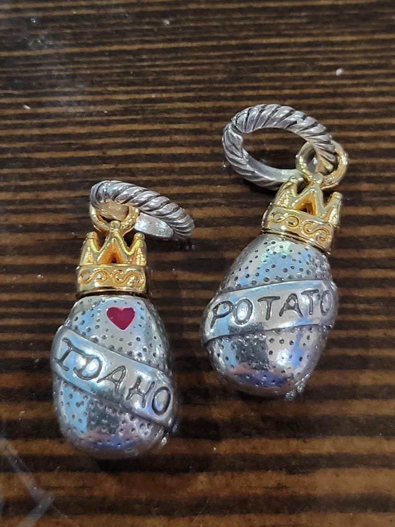 Puede incluir: Dos colgantes plateados con forma de patata y coronas doradas. Uno tiene grabado "IDAHO" con un coraz&oacute;n rojo, y el otro "POTATO". Ambos colgantes tienen un anillo plateado en la parte superior.