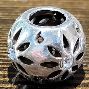 May include: A silver-colored, spherical bead with openwork floral designs. The bead features clear, round crystals embedded in the design. The bead is likely used for jewelry making or as a decorative element.