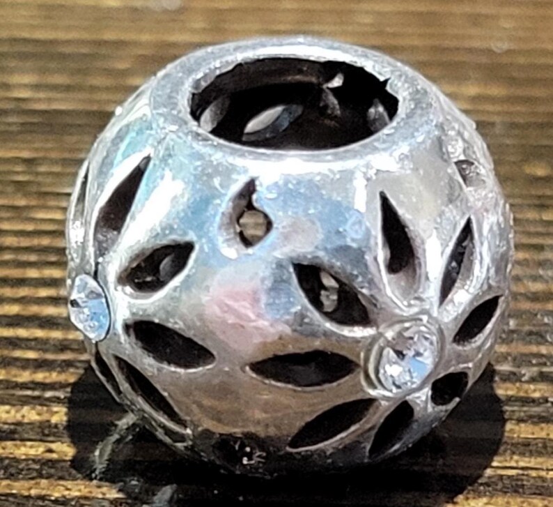May include: A silver-colored, spherical bead with openwork floral designs. The bead features clear, round crystals embedded in the design. The bead is likely used for jewelry making or as a decorative element.