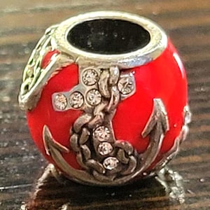 May include: A red bead with a silver anchor and chain design. The bead has a silver top and is embellished with clear crystals. The anchor and chain are detailed with silver accents. The bead is likely for a bracelet or necklace.