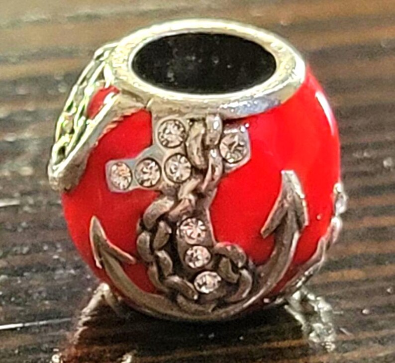 May include: A red bead with a silver anchor and chain design. The bead has a silver top and is embellished with clear crystals. The anchor and chain are detailed with silver accents. The bead is likely for a bracelet or necklace.