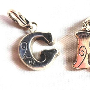 May include: Four silver-toned alphabet charms, spelling out the letters "F", "G", "H", and "I". Each charm has a decorative design and a clasp for attaching to a bracelet or necklace. The letters are in a serif font.
