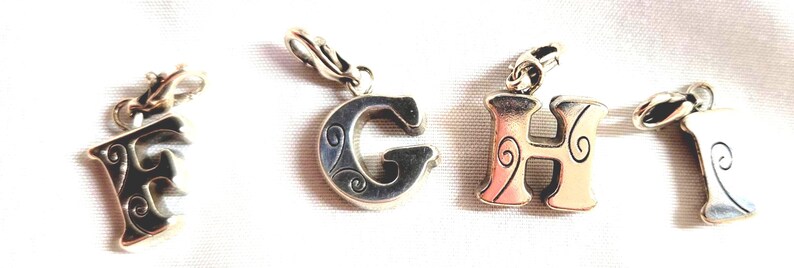 May include: Four silver-toned alphabet charms, spelling out the letters "F", "G", "H", and "I". Each charm has a decorative design and a clasp for attaching to a bracelet or necklace. The letters are in a serif font.