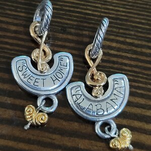 Puede incluir: Un par de pendientes plateados y dorados. Cada pendiente presenta un aro plateado, un dise&ntilde;o de nota musical dorada y un semic&iacute;rculo plateado con las palabras "SWEET HOME" y "ALABAMA" en negro. Un dije dorado cuelga debajo.