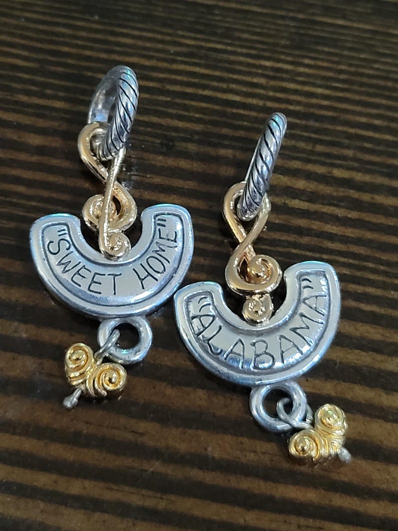 Puede incluir: Un par de pendientes plateados y dorados. Cada pendiente presenta un aro plateado, un dise&ntilde;o de nota musical dorada y un semic&iacute;rculo plateado con las palabras "SWEET HOME" y "ALABAMA" en negro. Un dije dorado cuelga debajo.