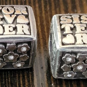 May include: Two silver-toned rings. One ring reads "FOREVER" with a heart, the other reads "SISTERS" with a heart. Each ring has a band of flower-shaped embellishments with a central gem.