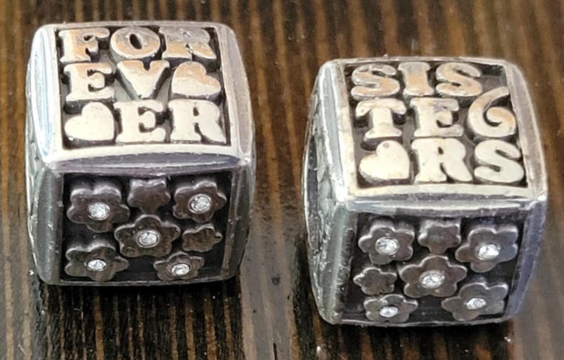 May include: Two silver-toned rings. One ring reads "FOREVER" with a heart, the other reads "SISTERS" with a heart. Each ring has a band of flower-shaped embellishments with a central gem.