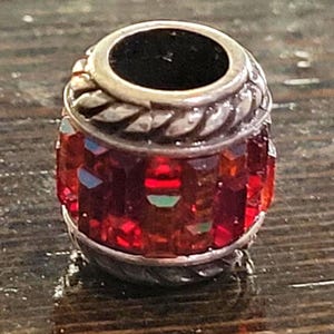 May include: A silver-toned bead with a cylindrical shape. The bead features a band of red, faceted crystals around the center. The top and bottom have a silver rope-like design. The top has a dark, circular opening.