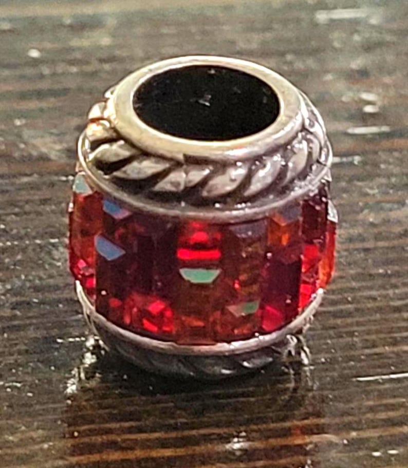 May include: A silver-toned bead with a cylindrical shape. The bead features a band of red, faceted crystals around the center. The top and bottom have a silver rope-like design. The top has a dark, circular opening.