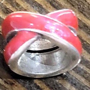 May include: A red and silver bead with a crisscross design. The bead has a glossy red enamel finish with silver-colored metal accents. The bead is cylindrical with a hole running through the center.