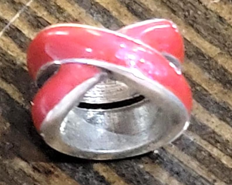 May include: A red and silver bead with a crisscross design. The bead has a glossy red enamel finish with silver-colored metal accents. The bead is cylindrical with a hole running through the center.