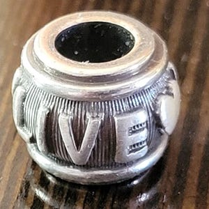 May include: Silver-toned charm with the word "LIVE" engraved on its surface. The charm has a cylindrical shape with a central hole and ribbed detailing. The letters are raised and stand out against the textured background.