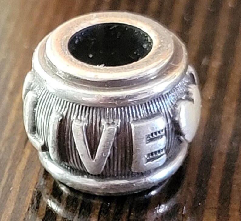 May include: Silver-toned charm with the word "LIVE" engraved on its surface. The charm has a cylindrical shape with a central hole and ribbed detailing. The letters are raised and stand out against the textured background.