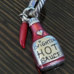 Puede incluir: Un charm plateado que representa una botella de salsa picante roja con el texto "Brighton HOT SAUCE" y un coraz&oacute;n. Un pimiento rojo cuelga junto a la botella. El charm tiene un lazo plateado retorcido.