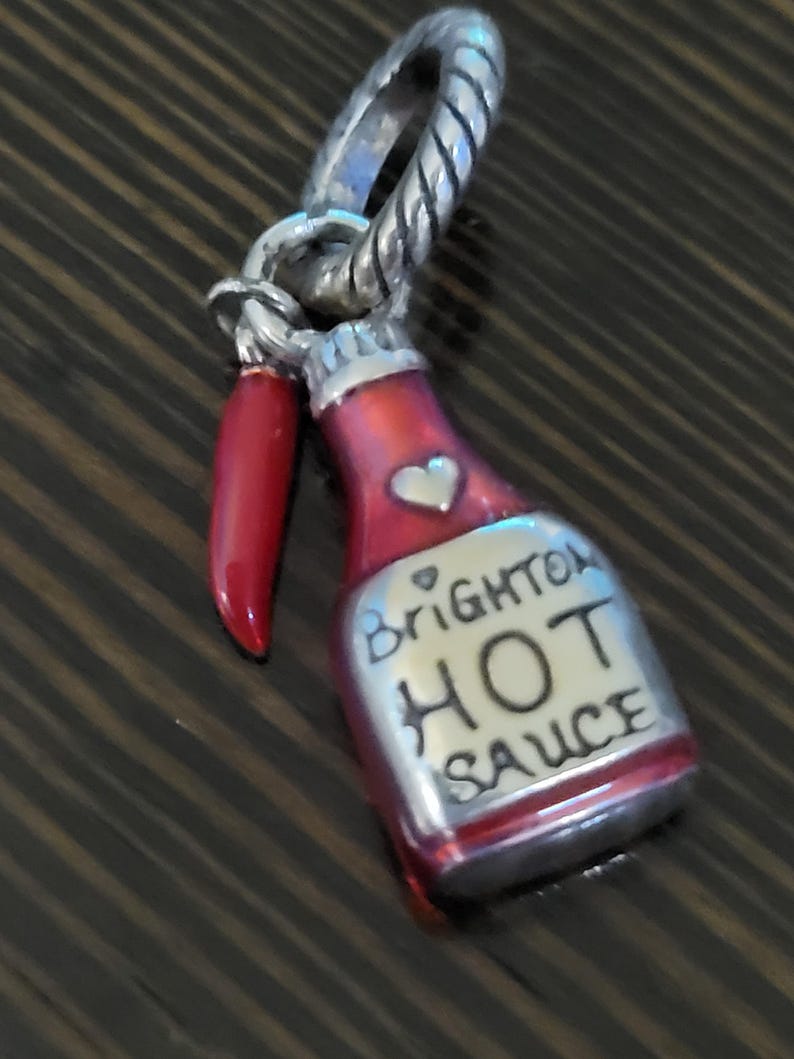 Puede incluir: Un charm plateado que representa una botella de salsa picante roja con el texto "Brighton HOT SAUCE" y un coraz&oacute;n. Un pimiento rojo cuelga junto a la botella. El charm tiene un lazo plateado retorcido.