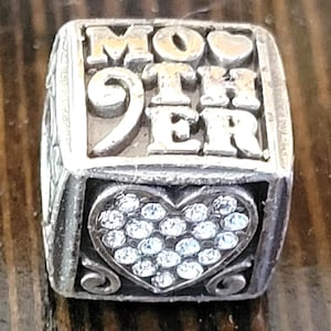 Op de afbeelding: Zilveren bedel met het woord "MOTHER" in verhoogde letters. Het nummer "9" is ook zichtbaar. Een hartvorm gevuld met kleine, heldere stenen staat op de voorkant. De bedel ligt op een houten oppervlak.