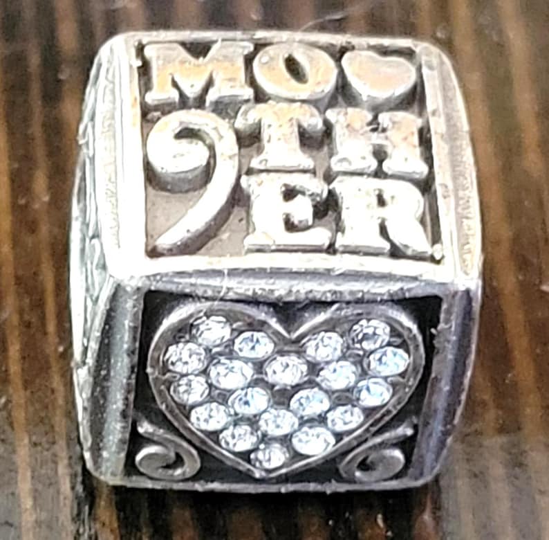 May include: Silver charm with the word "MOTHER" in raised letters. The number "9" is also visible. A heart shape filled with small, clear stones is on the front. The charm is on a wooden surface.