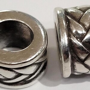 May include: Two silver-colored beads with a braided design. The beads have a cylindrical shape with a large hole running through the center. The braided pattern wraps around the bead's body, creating a textured surface.