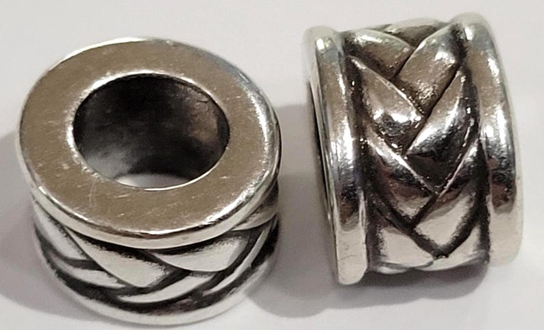 May include: Two silver-colored beads with a braided design. The beads have a cylindrical shape with a large hole running through the center. The braided pattern wraps around the bead's body, creating a textured surface.